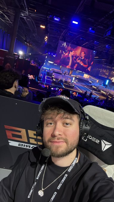 A young man wearing a black cap and headset takes a selfie in the foreground, with the Esports World Cup stage visible behind him, featuring a large screen showing players competing. Text visible in the image includes "EMCE" on his lanyard and shirt, "Paris" signage, and "S25" branding, aligning with the "Cast EWC Day 2" post highlighting his role in casting the Esports World Cup event.
