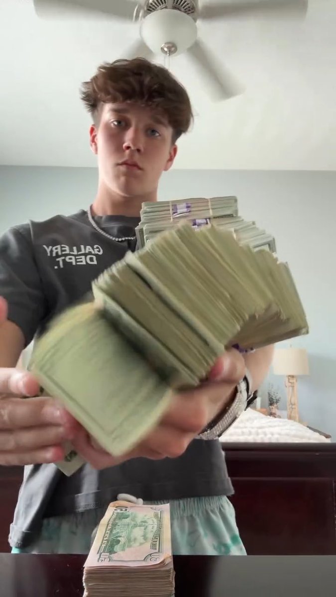 A teenage boy holding large stacks of US dollar bills toward the camera, wearing a dark t-shirt that reads 'GALLERY DEPT.' backwards, with another stack of money resting on a table in the foreground.