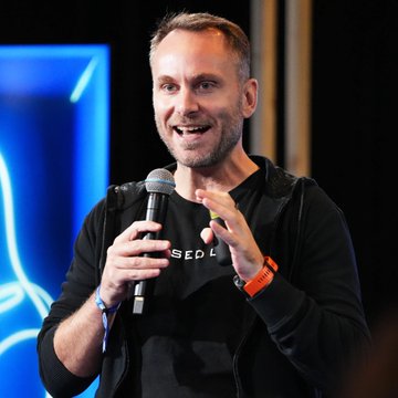 A man with a beard wearing a black sweater holds a microphone while speaking at what appears to be a conference or event.