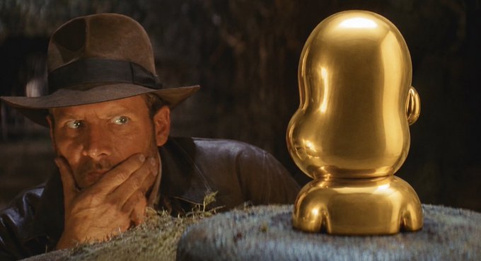 A cinematic still shows actor Harrison Ford as Indiana Jones in a contemplative pose next to a polished golden idol.