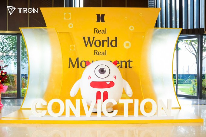 The image shows a TRON-branded promotional display at the Conviction 2026 event, featuring a large white one-eyed monster mascot and the text "Real World Real Movement" on a yellow backdrop, with "CONVICTION" spelled out in large white letters. The TRON logo appears in the top-left corner, visually confirming TRON's role as the Diamond Sponsor mentioned in the accompanying post about the Web3 event.