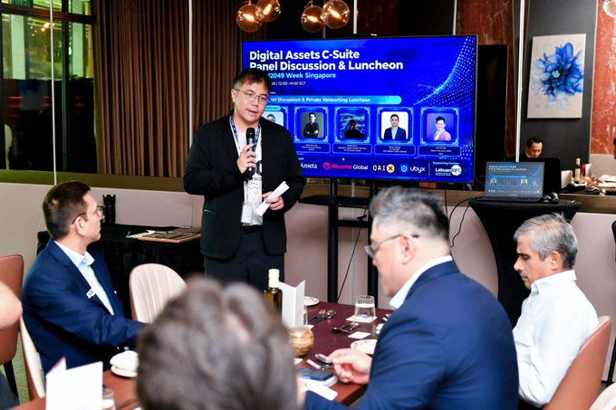 A man in a suit speaks at a "Digital Assets C-Suite Panel Discussion & Luncheon" event in Singapore (2049 Week Singapore, 16 Nov 19:30 SGT) with attendees seated at a round table.