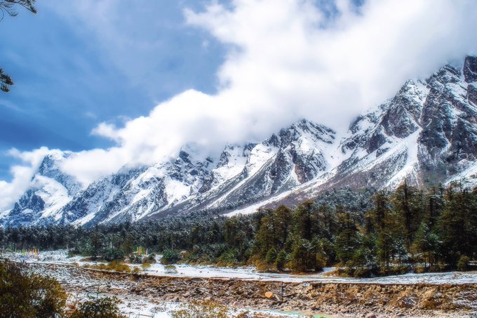 A scenic landscape view of snow-capped mountains rising above a forested valley under a sky filled with white clouds.