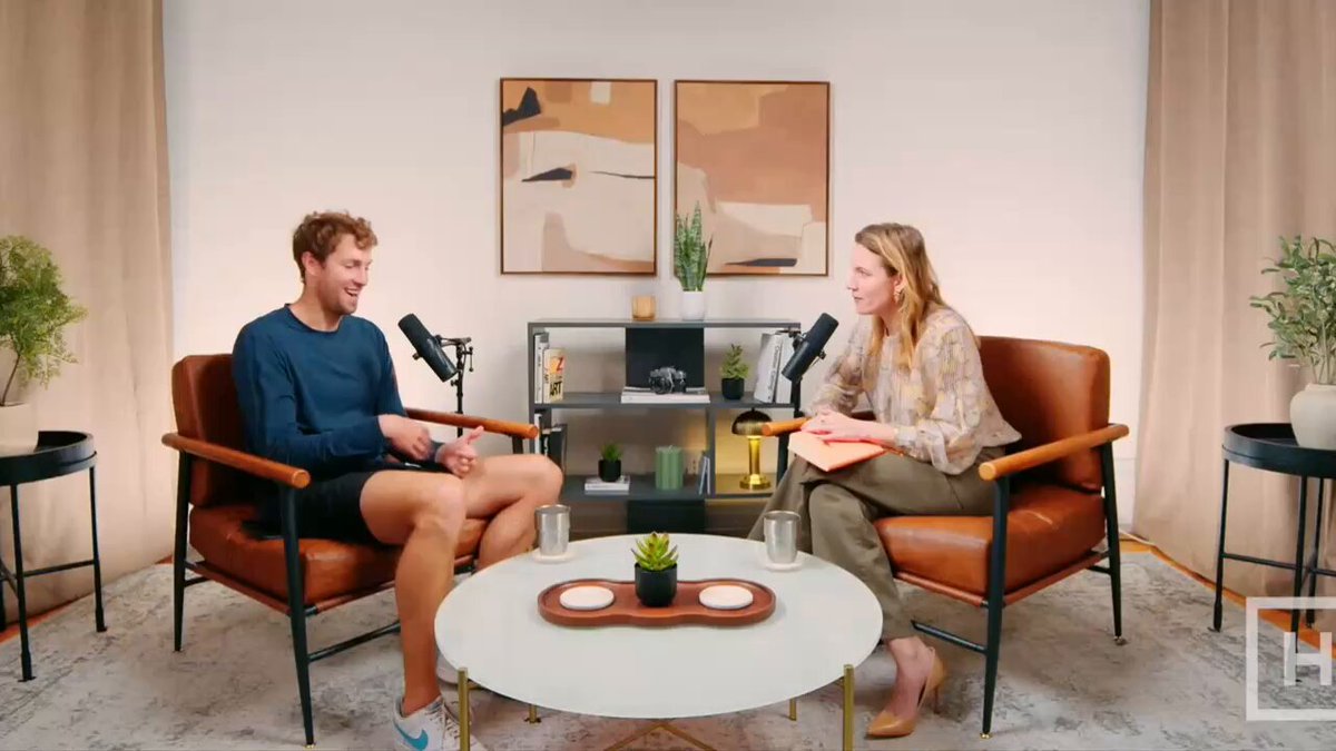 The image shows a man and woman sitting across from each other, engaged in conversation while seated in chairs. They are positioned in front of a coffee table with two cups placed on top of it. The room has white walls and beige carpeting, creating a cozy atmosphere for their discussion. A potted plant is visible to the left side of the image, adding a touch of greenery to the space.