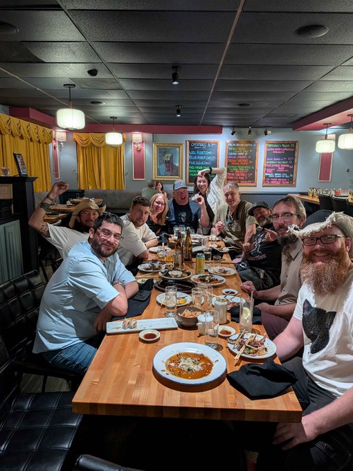 A large group of friends gather around a long wooden table at what appears to be the HOSKY unofficial after-party following RareEvo, with several attendees striking playful poses for the camera. The table is filled with half-eaten plates of pasta, drinks, and bottle caps, while chalkboards in the background display a menu featuring items like "Hosky Specialty Pizza" and prices ranging from $1.95 to $15.