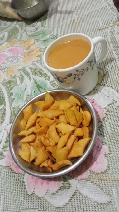 A traditional Indian breakfast setup featuring a white floral mug filled with masala chai alongside a metal plate of golden fried snacks on a floral patterned tablecloth.