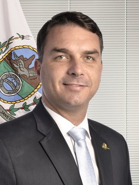 The image shows a man in a dark suit with a light blue tie, wearing a lapel pin, posed in front of what appears to be a Brazilian government or official seal. While the post references Flávio Bolsonaro and current Brazilian presidential election odds, the image does not visibly contain any text, numbers, or names identifying the individual, so any connection to Flávio Bolsonaro cannot be confirmed from the picture itself.