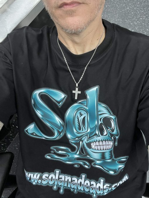 A man wearing a black t-shirt with a blue skull graphic spelling 'sol' and 'www.solanadeads.com' printed below, along with a silver cross necklace.