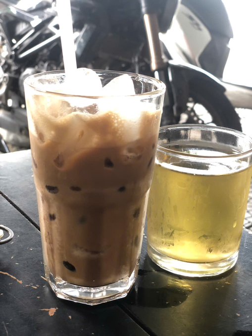 A tall glass of iced Thai milk tea next to a glass of green tea on a dark wooden table with a motorcycle in the background.