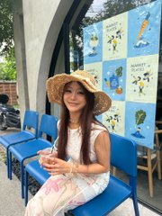 A woman wearing a straw sun hat and white sleeveless top sitting on blue chairs in what appears to be an outdoor event venue with colorful artwork displays behind her.
