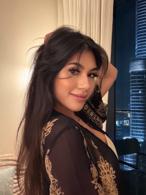 A woman with long dark hair poses for a selfie indoors, with a tall illuminated skyscraper visible through the window behind her.