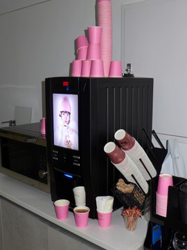 An espresso machine decorated with stacks of pink paper cups, featuring a digital screen displaying an image of a person wearing glasses, with coffee supplies, stirrers, and additional cups arranged on the counter.