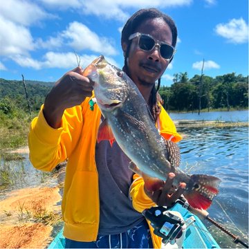 A man wearing sunglasses and a yellow hoodie smiles while holding up a large peacock bass fish he caught, standing on a small boat on a lake.