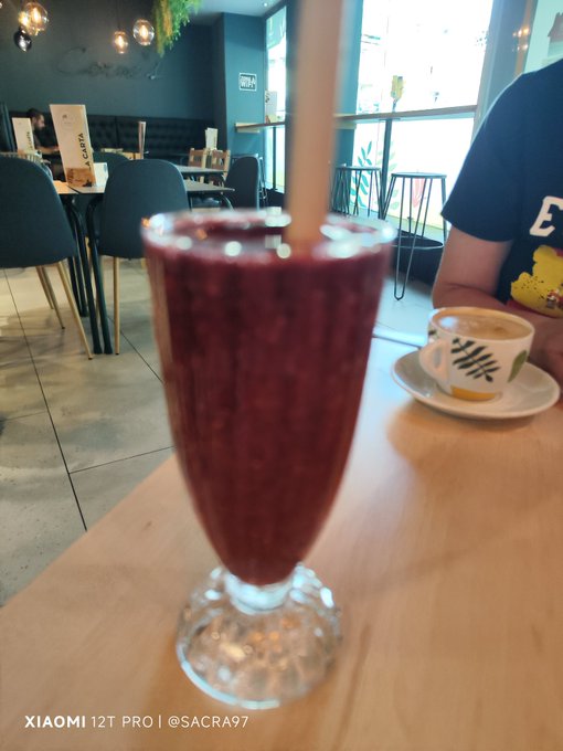A tall glass of a dark red/purple smoothie with a straw sits on a cafe table next to a coffee cup, with a person seated across in the background.