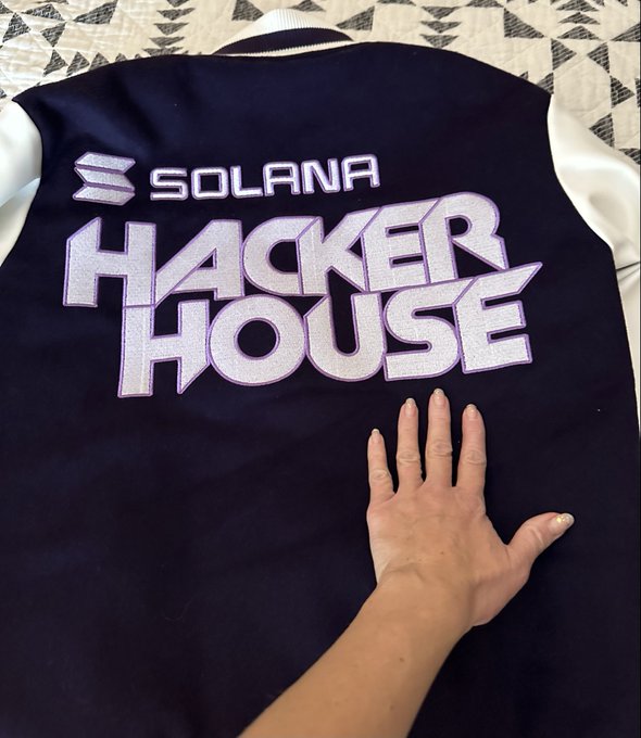 A dark navy varsity-style jacket with white sleeves displays an embroidered Solana logo and large 'HACKER HOUSE' text in white and lavender, with a hand resting on the front.