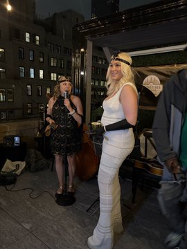 Two women in 1920s-style flapper attire—one in a black dress with a headband, the other in a white outfit with a black belt—pose on a rooftop stage beside an upright bass and a sign reading "THE BITCOIN BAR," capturing the Gatsby-esque vibe of the meetup highlighted in the post.