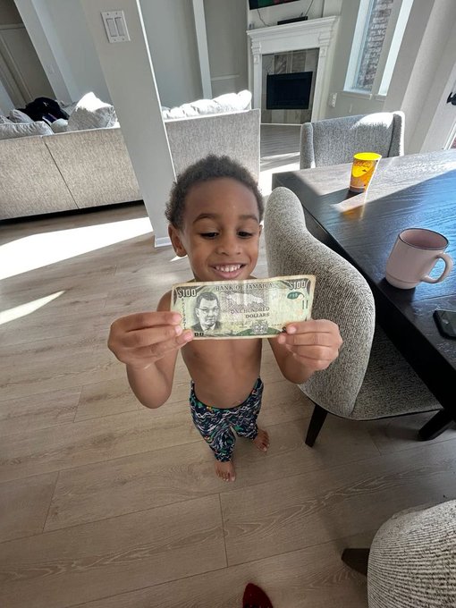 A young child in a diaper smiling and holding up a Jamaican $100 banknote in a home setting with a dining table and chairs visible.