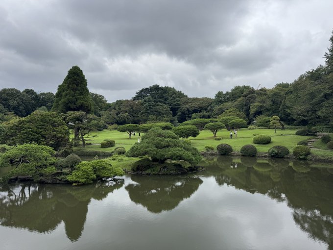 A serene Japanese garden featuring manicured pine trees and shrubs beside a calm reflecting pond under an overcast sky, with two small figures visible in the distance.
