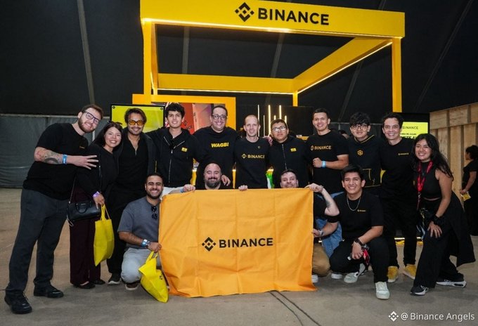 The Binance Angels team poses together with a large Binance-branded yellow flag at the Fin & Fun Fest event in Lima, Peru, celebrating the crypto community gathering. The group of staff members, all wearing matching black Binance apparel, smile for a group photo in front of the company's distinctive yellow and black booth, perfectly capturing the spirit of connection and community highlighted in the post.
