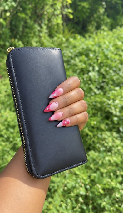 A hand with long pink and white decorated nails holds a black leather zip-around wallet against a background of green foliage.