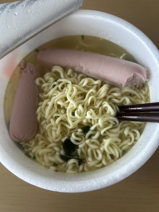 An overhead view of an open styrofoam cup containing instant ramen noodles in broth, topped with two pink sausages, with dark chopsticks resting on the side.