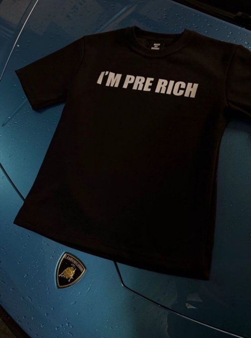 A black t-shirt with 'I'M PRE RICH' printed in white lies on a wet blue car hood, with a Lamborghini logo badge beside it.