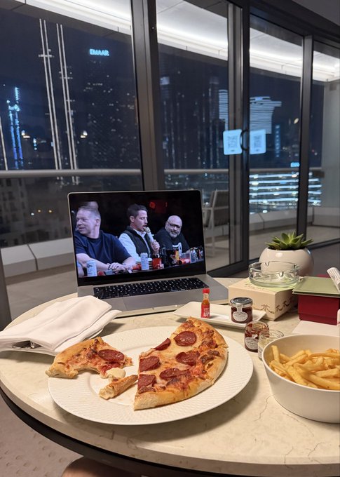 A table set with pizza, fries, and condiments in front of an open laptop displaying a podcast or video with three men, overlooking a nighttime city skyline through floor-to-ceiling windows.