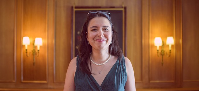 A smiling woman with sunglasses on her head and a pearl necklace stands in an elegantly wood-paneled room with warm wall sconces.