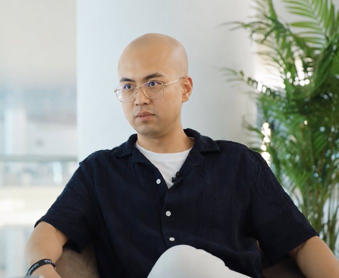 The image features a man with a shaved head wearing glasses sitting in a chair. He appears to be looking off into the distance or deep in thought. The background of the image includes a plant, suggesting an indoor setting such as a living room or office space. There is also text present that reads "Focus on what is in the image and how it relates to the post".