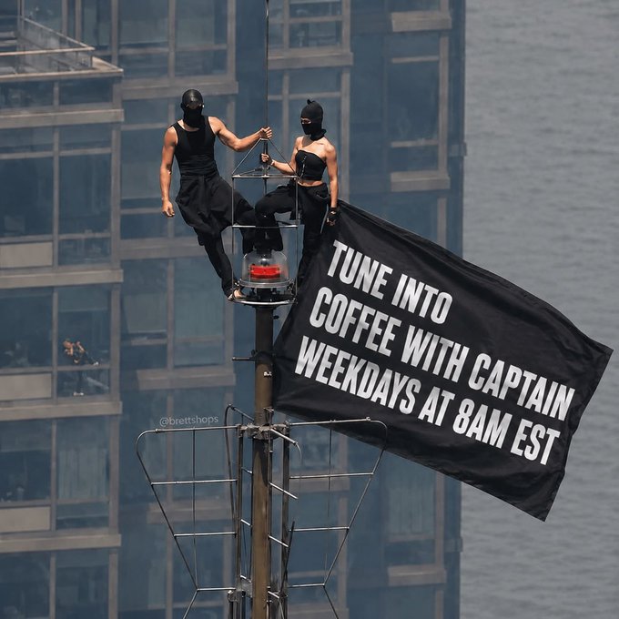 Two performers in black balaclavas and dark clothing standing on a tall pole/stunt structure, one holding a large black banner reading 'TUNE INTO COFFEE WITH CAPTAIN WEEKDAYS AT 8AM EST.'