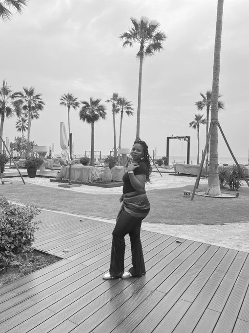 A woman poses confidently on a wooden deck at a tropical beach resort, surrounded by tall palm trees and ocean views, radiating a joyful, victorious energy that matches her uplifting "GM" morning affirmation of triumph and gratitude.