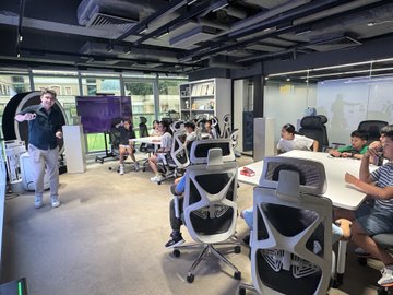 An open-plan office space with exposed ceiling ductwork features several people working at desks with white office chairs, while one man stands on the left gesturing.