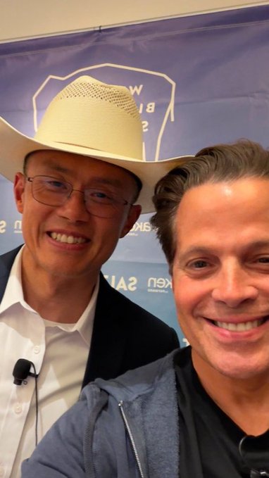A selfie of two smiling men at an event, one wearing a white cowboy hat and glasses, with a purple backdrop partially reading 'A21' and 'Kex'.