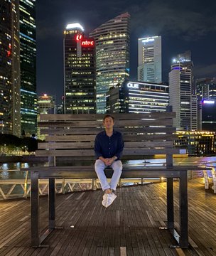 A young man in a navy shirt and white pants sitting on a wooden bench at night, with the illuminated Singapore skyline featuring the NTUC building visible across the water behind him.