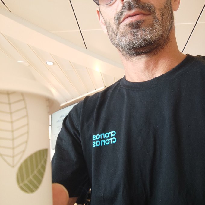 A close-up photograph of a bearded man wearing a black t-shirt with cyan text that reads 'SOTOS SOTOS' backwards. He appears to be holding a white paper cup decorated with leaf patterns in the foreground, with a ceiling visible above him.