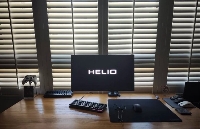 A minimalist wooden desk setup features a central monitor displaying the word 'HELIO', accompanied by a mechanical keyboard, mouse, game controller on a stand, and scissors, all set against a backdrop of closed horizontal window blinds.
