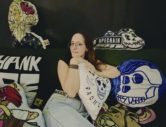 A young woman with glasses poses among skull-themed plush toys and pillows, some featuring 'APECHAIN' branding and stylized skeletal artwork.