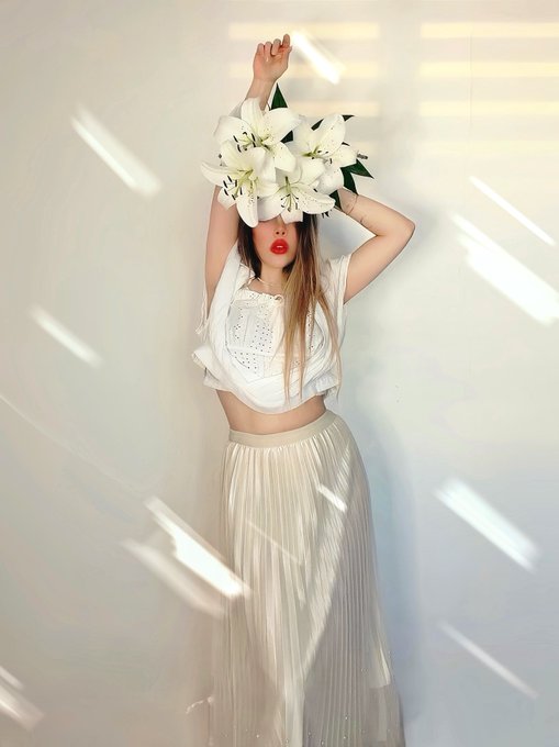 A fashion editorial photo of a woman in a white crop top and pleated skirt holding large white lilies in front of her face, standing against a white wall with diagonal light streaks.