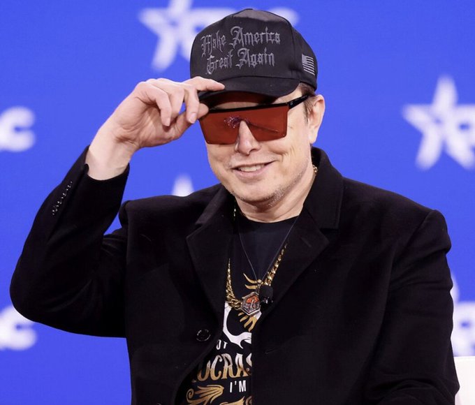 A man smiles while lifting a pair of red-tinted sunglasses, wearing a black cap reading 'Make America Great Again,' a black blazer, and a graphic t-shirt with gold chains, set against a blue background with white stars.