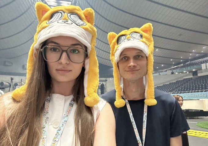 Photo of a young woman with glasses and a young man wearing matching yellow cat/pilot hats at what appears to be a university event or conference.