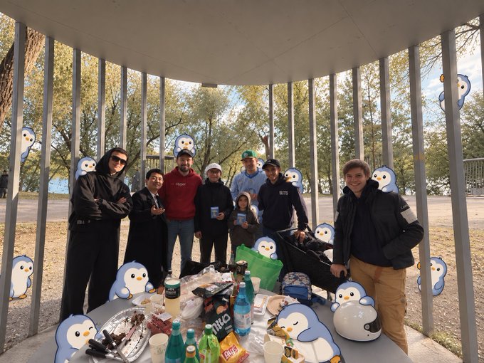 Group of friends gathered under a circular pavilion for a picnic with drinks and snacks, decorated with many penguin stickers overlaid on the image.