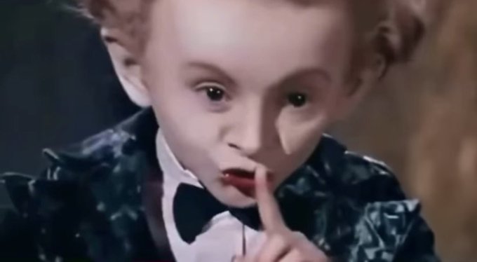A vintage cinematic close-up of a young child in a dark ruffled outfit and bow tie holding a finger to their lips in a shushing gesture.