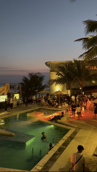 An evening photo captures a lively beach club or pool party scene at dusk, with people socializing around a green-lit swimming pool near the ocean, featuring palm trees and a building with a "PINA" sign.