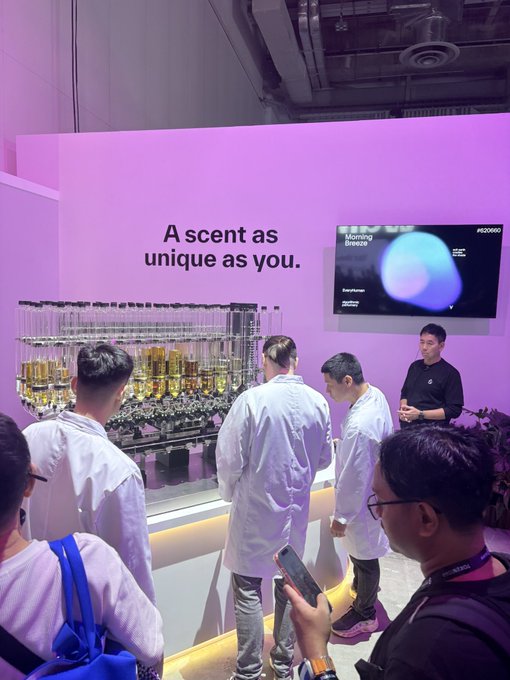 An exhibition booth with a pink wall reading 'A scent as unique as you.' showing a display of perfume bottles and a screen labeled 'Morning Breeze' with people in lab coats observing.