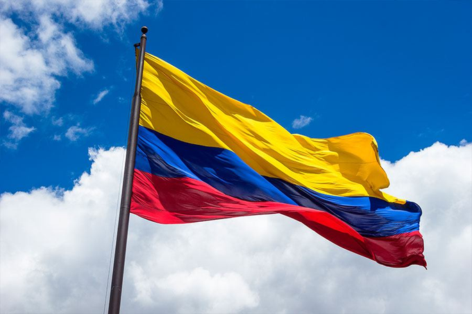 A Colombian flag waves against a bright blue sky, symbolizing Colombia's growing institutional adoption of Bitcoin as the country's second-largest pension fund allocates to the cryptocurrency. The image visually ties to the post's message about emerging market institutions embracing BTC as part of the global financial landscape.