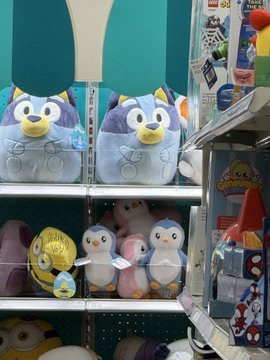 Plush toys of penguins and other animals are displayed on store shelves, including two prominent gray penguin plushies on the top shelf. The post highlights the Pudgy Penguins merchandise as a retail expansion, referencing the $PENGU ticker.