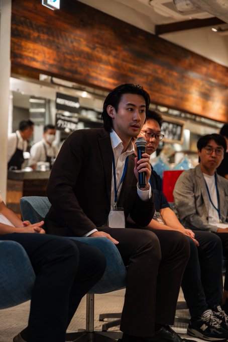 An Asian man in a dark suit and white shirt holding a microphone while speaking at what appears to be a panel discussion or business event.