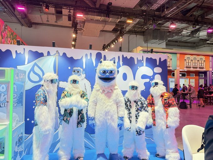 Five people in white furry yeti costumes with sunglasses and colorful shirts pose together at what appears to be a brand event or convention.