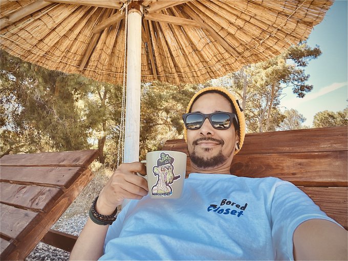 A man wearing sunglasses and a yellow beanie relaxes under a thatched umbrella, holding a coffee mug while wearing a shirt that reads "Bored Closet," tying into the "GM web3" post by embodying a chill, leisurely vibe typical of crypto community greetings.