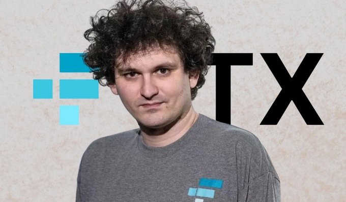 A young man with curly hair wearing a gray t-shirt poses in front of a backdrop displaying a blue logo and the black letters 'TX'.